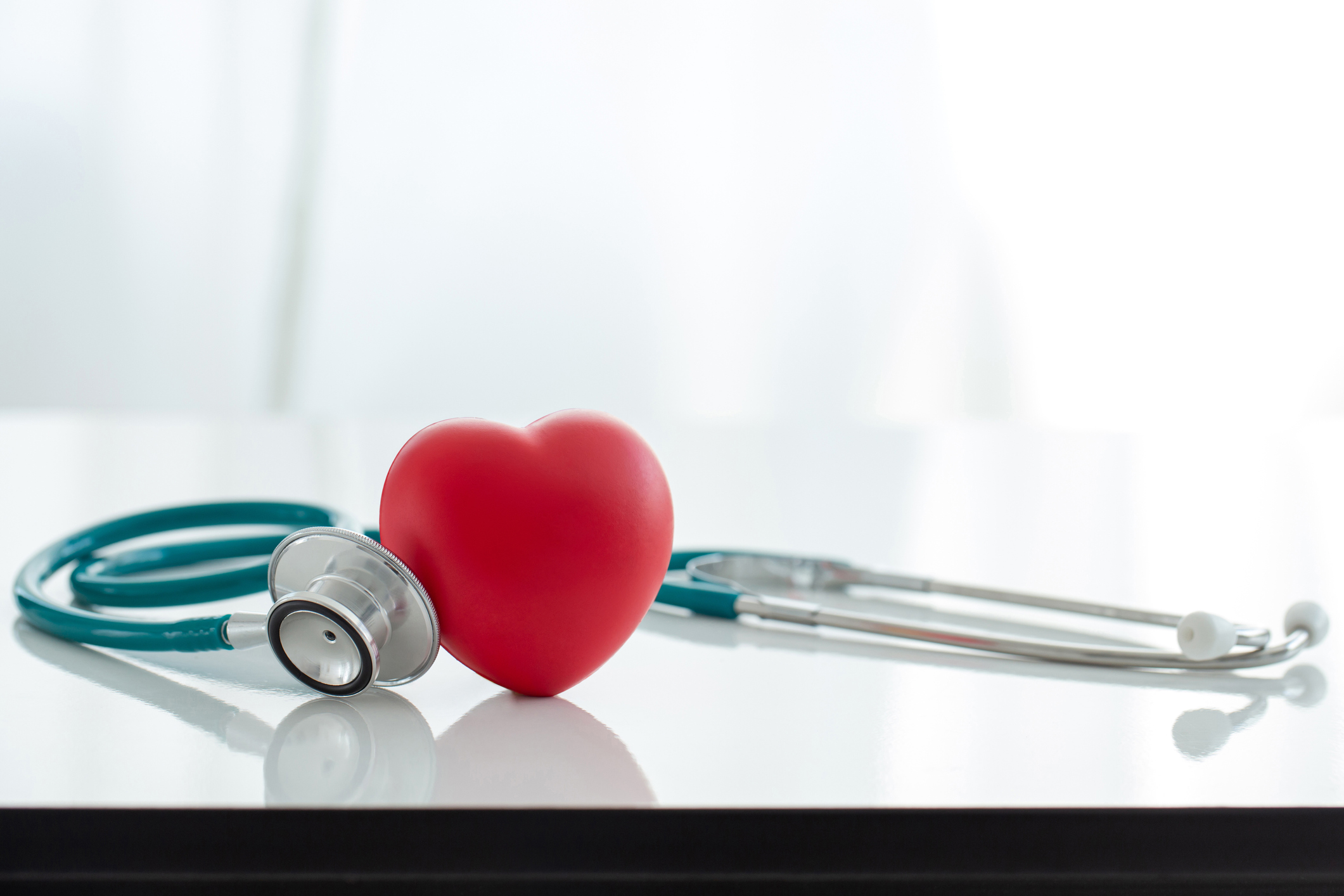 A blue and silver stethoscope and a red heart-shaped stress ball on a white table with a bright white background. 